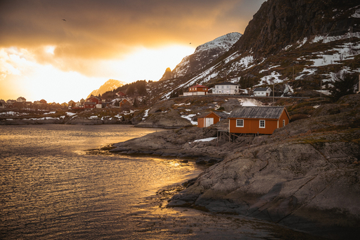 View of the orange houses nestled on rocky shores their windows glowing warmly against the backdrop of snow dusted mountains and golden sunset Tind Nordland Norway.