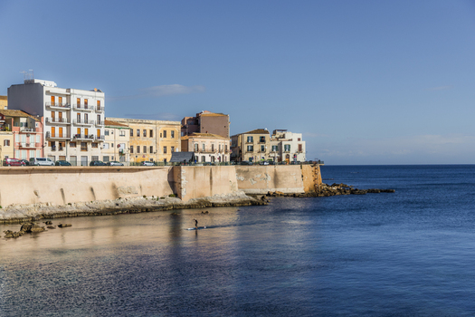 View of buildings with a stone wall along the coast meeting the serene blue sea under a clear sky Ortigia Sicily Italy.