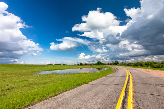 View of a rural road with yellow lane markings curving through a green field with a puddle under a dramatic sky with heavy storm clouds in Oklahoma City Oklahoma United States.