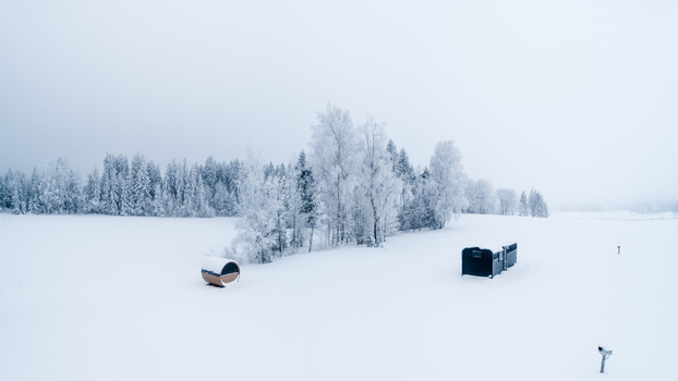 Aerial view of a barrel sauna and a small structure in a vast snow covered landscape with frost covered trees under a bright overcast sky in Latvia. Aerial view of a barrel sauna and a small structure in a vast snow covered landscape with frost covered trees under a bright overcast sky in Latvia.