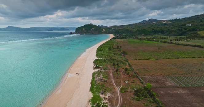 Aerial view of the turquoise waters and sandy coastline of Lombok featuring lush green hills and agricultural fields under a cloudy sky in Lombok West Nusa Tenggara Indonesia.