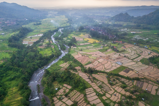 Aerial view of lush terraced rice fields and a winding river flowing through a tropical valley under a misty sky in Sidemen Bali Indonesia.