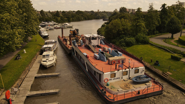 Aerial view of the Joel Leclercq barge navigating a canal alongside moored pleasure boats surrounded by lush green banks and trees in Saint Omer Hauts de France France. Aerial view of the Joel Leclercq barge navigating a canal alongside moored pleasure boats surrounded by lush green banks and trees in Saint Omer Hauts de France France.