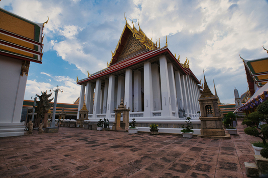 View of The Grand Palace with its ornate golden roof and white columns set against a cloudy sky in a paved courtyard in Bangkok Thailand. View of The Grand Palace with its ornate golden roof and white columns set against a cloudy sky in a paved courtyard in Bangkok Thailand.