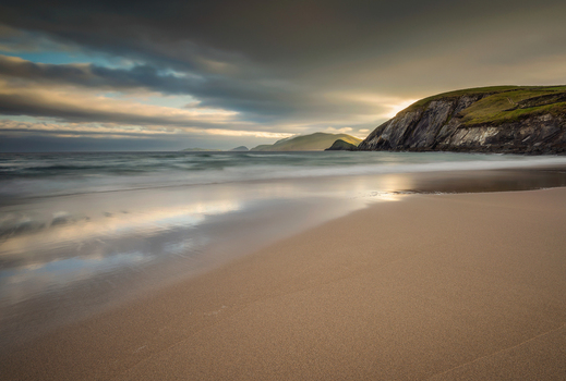 View of the sandy shore at Dunmore Head with dramatic cliffs and ocean waves under a cloudy sky in Ireland.