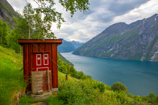 View of a small red wooden outhouse with heart shaped cutouts at the historic Smage mountain farm overlooking the Geirangerfjord and steep mountains in Geiranger More og Romsdal Norway.