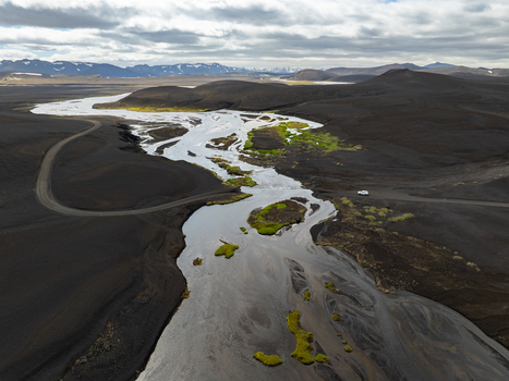 Aerial view of a winding river flowing through a vast dark volcanic landscape with patches of green moss and distant snow capped mountains in Iceland. Aerial view of a winding river flowing through a vast dark volcanic landscape with patches of green moss and distant snow capped mountains in Iceland.