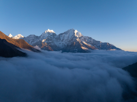 Aerial view of snow capped Himalayan mountain peaks rising above a dense sea of clouds under a clear blue sky in Nepal.