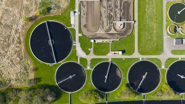 Aerial view of the RWZI Geestmerambacht wastewater treatment plant featuring circular clarifiers and rectangular aeration tanks surrounded by green lawns and trees. Aerial view of the RWZI Geestmerambacht wastewater treatment plant featuring circular clarifiers and rectangular aeration tanks surrounded by green lawns and trees.