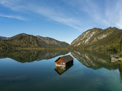 Aerial view of a wooden boathouse floating on the calm clear water of a lake surrounded by forested mountains under a bright blue sky in Rottenbach Bayern Germany. Aerial view of a wooden boathouse floating on the calm clear water of a lake surrounded by forested mountains under a bright blue sky in Rottenbach Bayern Germany.