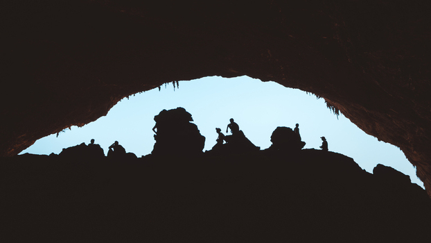 View of silhouetted people standing inside the large entrance of Hoq Cave with rocky formations and bright sky visible in Socotra Socotra Yemen. View of silhouetted people standing inside the large entrance of Hoq Cave with rocky formations and bright sky visible in Socotra Socotra Yemen.