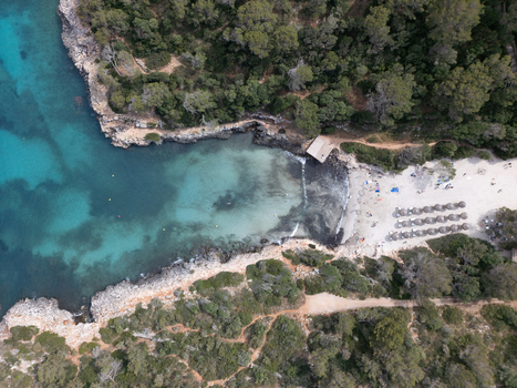 Aerial view of Cala Sa Nau turquoise waters rocky cliffs and a sandy beach with sunbeds surrounded by lush pine forests in Felanitx Balearic Islands Spain. Aerial view of Cala Sa Nau turquoise waters rocky cliffs and a sandy beach with sunbeds surrounded by lush pine forests in Felanitx Balearic Islands Spain.
