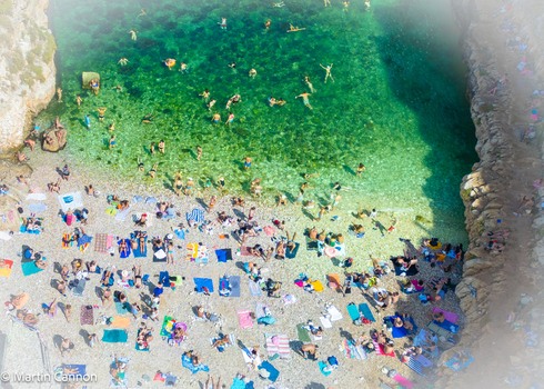 Aerial view of the popular Lama Monachile beach with people swimming in turquoise water and sunbathing on the pebble shore surrounded by rocky cliffs in Polignano a Mare Apulia Italy.