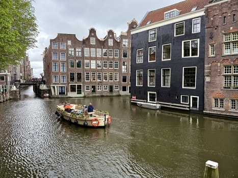 View of a boat with people cruising along the Amsterdam canals surrounded by historic narrow houses with gabled facades in the Netherlands. View of a boat with people cruising along the Amsterdam canals surrounded by historic narrow houses with gabled facades in the Netherlands.