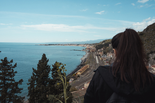 View of a woman with dark hair gazing out over the tranquil turquoise coastline and historic town nestled between the sea and mountains Taormina Sicily Italy. View of a woman with dark hair gazing out over the tranquil turquoise coastline and historic town nestled between the sea and mountains Taormina Sicily Italy.