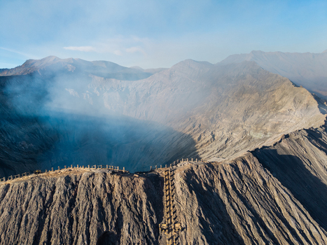 Aerial view of Mount Bromo crater with volcanic smoke rising from the caldera rugged rocky slopes and a wooden staircase leading to the rim in Tosari East Java Indonesia.