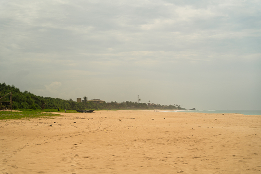 View of a wide sandy beach with a wooden boat lush green vegetation and distant buildings under an overcast sky in Sri Lanka. View of a wide sandy beach with a wooden boat lush green vegetation and distant buildings under an overcast sky in Sri Lanka.