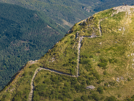 Aerial view of a winding stone hiking trail climbing a steep grassy mountain ridge with hikers standing on the summit above a vast forested valley in France.