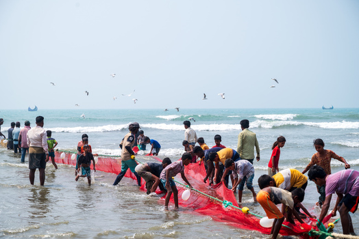 Teknaf Bangladesh 10 March 2026 View of fishermen hauling in a vibrant red net along the sandy shore as seabirds circle above the tranquil sea contrasting with the bright sky.