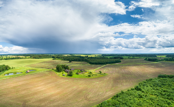 Aerial view of a rural landscape featuring a small farmhouse surrounded by cultivated fields green meadows and patches of forest under a dramatic cloudy sky Latvia. Aerial view of a rural landscape featuring a small farmhouse surrounded by cultivated fields green meadows and patches of forest under a dramatic cloudy sky Latvia.