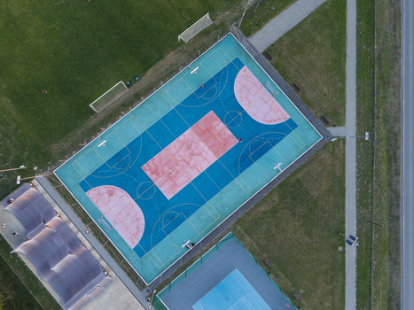 Aerial view of a vibrant multi colored outdoor sports court with basketball hoops surrounded by green grass and a nearby road in Poland.