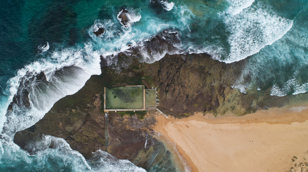 Aerial view of Newport Rock Pool with turquoise waves crashing against dark rocks and a sandy beach in Sydney New South Wales Australia.