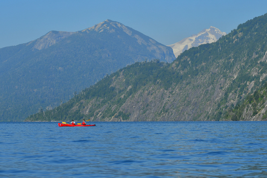 View of two people in a red kayak on the calm waters of Nahuel Huapi Lake with steep forested mountains and a clear blue sky in the background San Carlos de Bariloche Rio Negro Argentina. View of two people in a red kayak on the calm waters of Nahuel Huapi Lake with steep forested mountains and a clear blue sky in the background San Carlos de Bariloche Rio Negro Argentina.