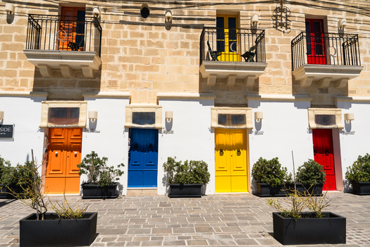 View of colorful traditional doors in orange blue yellow and red on a limestone building facade with wrought iron balconies in Marsaskala Southern Region Malta. View of colorful traditional doors in orange blue yellow and red on a limestone building facade with wrought iron balconies in Marsaskala Southern Region Malta.