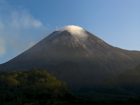 Aerial view of Mount Merapi volcano with steam rising from the crater above lush tropical forests under a clear blue sky Kecamatan Pakem Daerah Istimewa Yogya Indonesia. Aerial view of Mount Merapi volcano with steam rising from the crater above lush tropical forests under a clear blue sky Kecamatan Pakem Daerah Istimewa Yogya Indonesia.