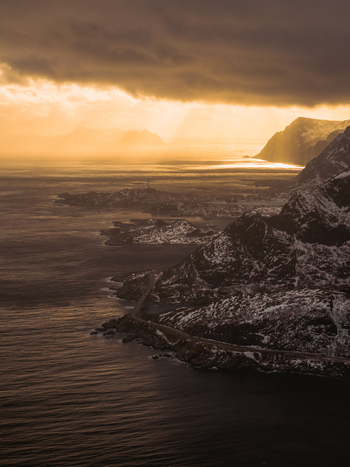 Aerial view of dramatic skies meet the rugged snow dusted peaks and dark rippling waters surrounding Reinebringen Nordland Norway.