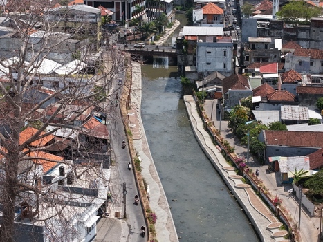 Aerial view of a canal flowing through the urban landscape with traditional rooftops a narrow road with motorcyclists and lush trees in Kecamatan Denpasar Barat Bali Indonesia. Aerial view of a canal flowing through the urban landscape with traditional rooftops a narrow road with motorcyclists and lush trees in Kecamatan Denpasar Barat Bali Indonesia.