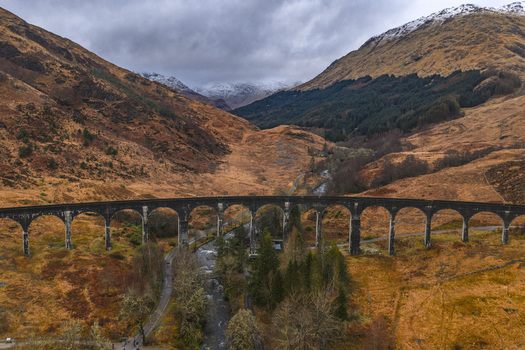 Aerial view of the historic Glenfinnan Viaduct with its iconic curved arches spanning a valley between autumn colored hills and snow capped mountains in Glenfinnan Scotland United Kingdom.