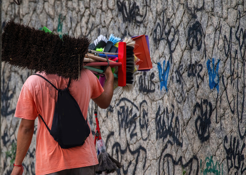 View of a street vendor carrying a large bundle of colorful brooms and cleaning tools on his shoulder past a graffiti covered stone wall in Rio de Janeiro State of Rio de Janeiro Brazil. View of a street vendor carrying a large bundle of colorful brooms and cleaning tools on his shoulder past a graffiti covered stone wall in Rio de Janeiro State of Rio de Janeiro Brazil.