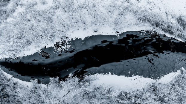 Aerial view of a dark winding river flowing through a snow covered forest in Latvia. Aerial view of a dark winding river flowing through a snow covered forest in Latvia.