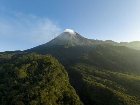Aerial view of Mount Merapi volcano with smoke rising from the crater above lush green forests under a clear blue sky in Kecamatan Pakem Daerah Istimewa Yogya Indonesia. Aerial view of Mount Merapi volcano with smoke rising from the crater above lush green forests under a clear blue sky in Kecamatan Pakem Daerah Istimewa Yogya Indonesia.