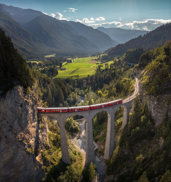 Aerial view of the Landwasser Viaduct with a red train crossing the stone arches set against alpine forests mountains and a river valley in Bergun Filisur Graubunden Switzerland.