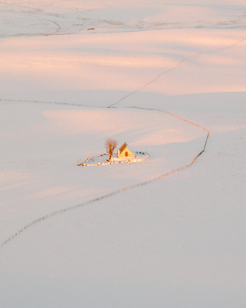 Aerial view of a golden lit building and tree stand out against the vast snow covered landscape illuminated by the soft glow of dawn Nasbinals Occitanie France.