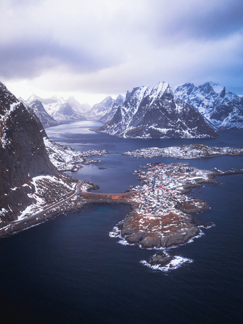 Aerial view of a picturesque village nestled among snow dusted islands and fjords framed by towering mountains under a moody sky Reinebringen Nordland Norway.