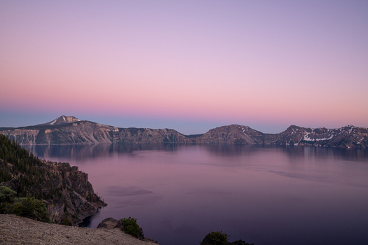 View of tranquil lake mirroring the soft pastel hues of the twilight sky framed by rugged cliffs and distant mountain peaks Crater Lake Oregon United States. View of tranquil lake mirroring the soft pastel hues of the twilight sky framed by rugged cliffs and distant mountain peaks Crater Lake Oregon United States.