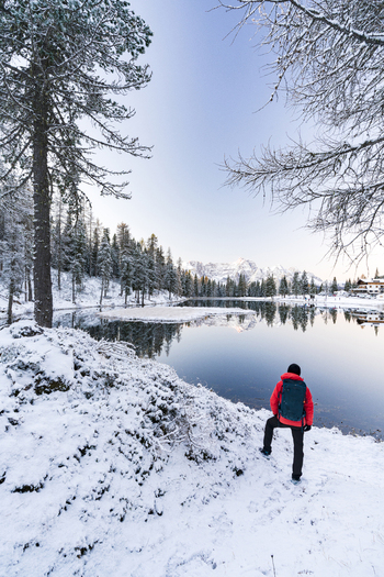View of a hiker standing near a tranquil lake with snow covered banks and a backdrop of majestic mountains Auronzo di Cadore Veneto Italy. View of a hiker standing near a tranquil lake with snow covered banks and a backdrop of majestic mountains Auronzo di Cadore Veneto Italy.