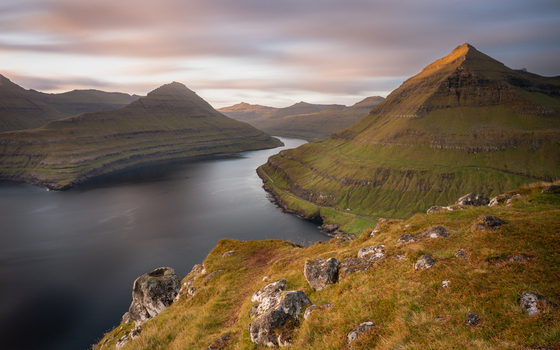 View of dramatic mountains and a deep fjord under a soft sunset sky in the Faroe Islands. View of dramatic mountains and a deep fjord under a soft sunset sky in the Faroe Islands.