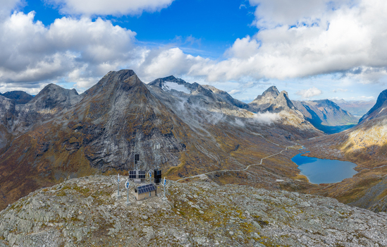 Aerial view of a mountain station with solar panels on a rocky peak overlooking a winding road and blue alpine lake surrounded by rugged mountains in Andalsnes More og Romsdal Norway.