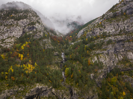 Aerial view of the Pyrenees mountains featuring a cascading waterfall amidst vibrant autumn foliage and rocky cliffs partially obscured by low hanging clouds in Spain. Aerial view of the Pyrenees mountains featuring a cascading waterfall amidst vibrant autumn foliage and rocky cliffs partially obscured by low hanging clouds in Spain.