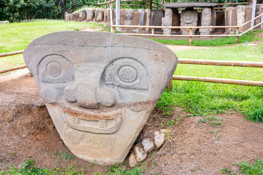 View of the ancient stone statue with a carved face at San Agustin Archaeological Park set in a grassy field with bamboo fencing in San Agustin Huila Colombia.