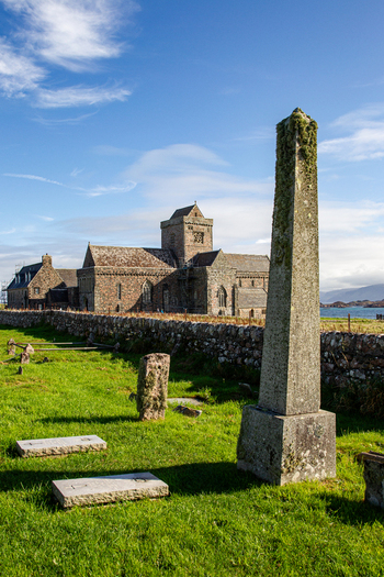 View of Iona Abbey and Nunnery with a historic stone monument and graveyard under a bright blue sky in Isle of Iona Scotland United Kingdom. View of Iona Abbey and Nunnery with a historic stone monument and graveyard under a bright blue sky in Isle of Iona Scotland United Kingdom.