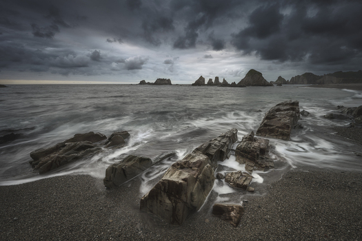 View of sharp rock formations on a pebble beach under a dramatic stormy sky with long exposure waves in Asturias Asturias Spain. View of sharp rock formations on a pebble beach under a dramatic stormy sky with long exposure waves in Asturias Asturias Spain.