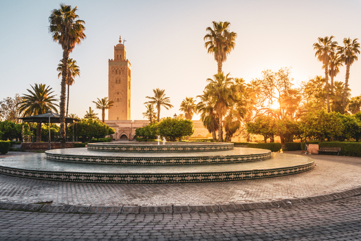 View of the Koutoubia Mosque with its minaret rising above a circular fountain surrounded by palm trees and greenery in the golden sunset light in Marrakesh Marrakesh Safi Morocco. View of the Koutoubia Mosque with its minaret rising above a circular fountain surrounded by palm trees and greenery in the golden sunset light in Marrakesh Marrakesh Safi Morocco.