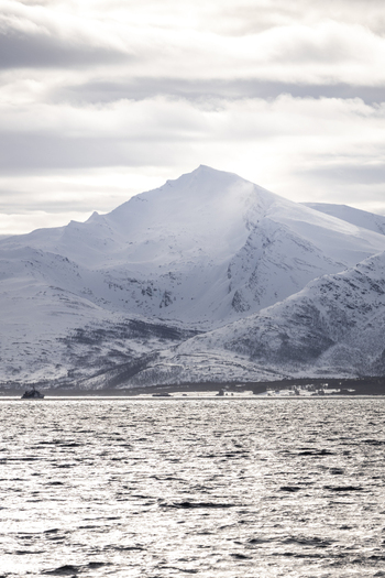 View of snow capped mountain peaks rising above the cold waters of a fjord with a small boat in the distance under a cloudy sky in Tromso Troms Norway.