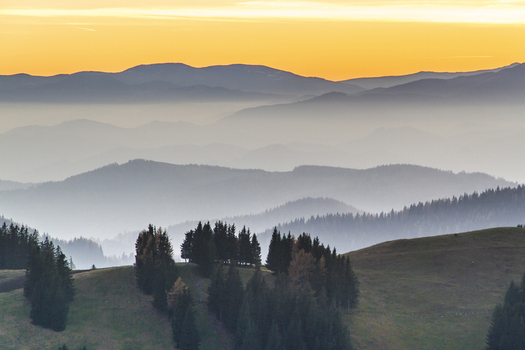 View of hazy mountain ranges fading into the distance under a golden sky with lush green hills in the foreground Sommeralm Steiermark Austria. View of hazy mountain ranges fading into the distance under a golden sky with lush green hills in the foreground Sommeralm Steiermark Austria.