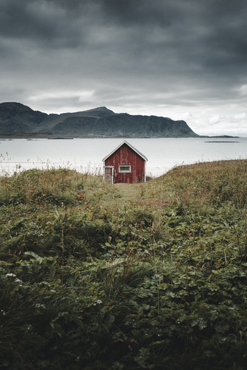 View of a traditional red wooden cabin set in a grassy field with mountains and water in the background in Ramberg Nordland Norway. View of a traditional red wooden cabin set in a grassy field with mountains and water in the background in Ramberg Nordland Norway.